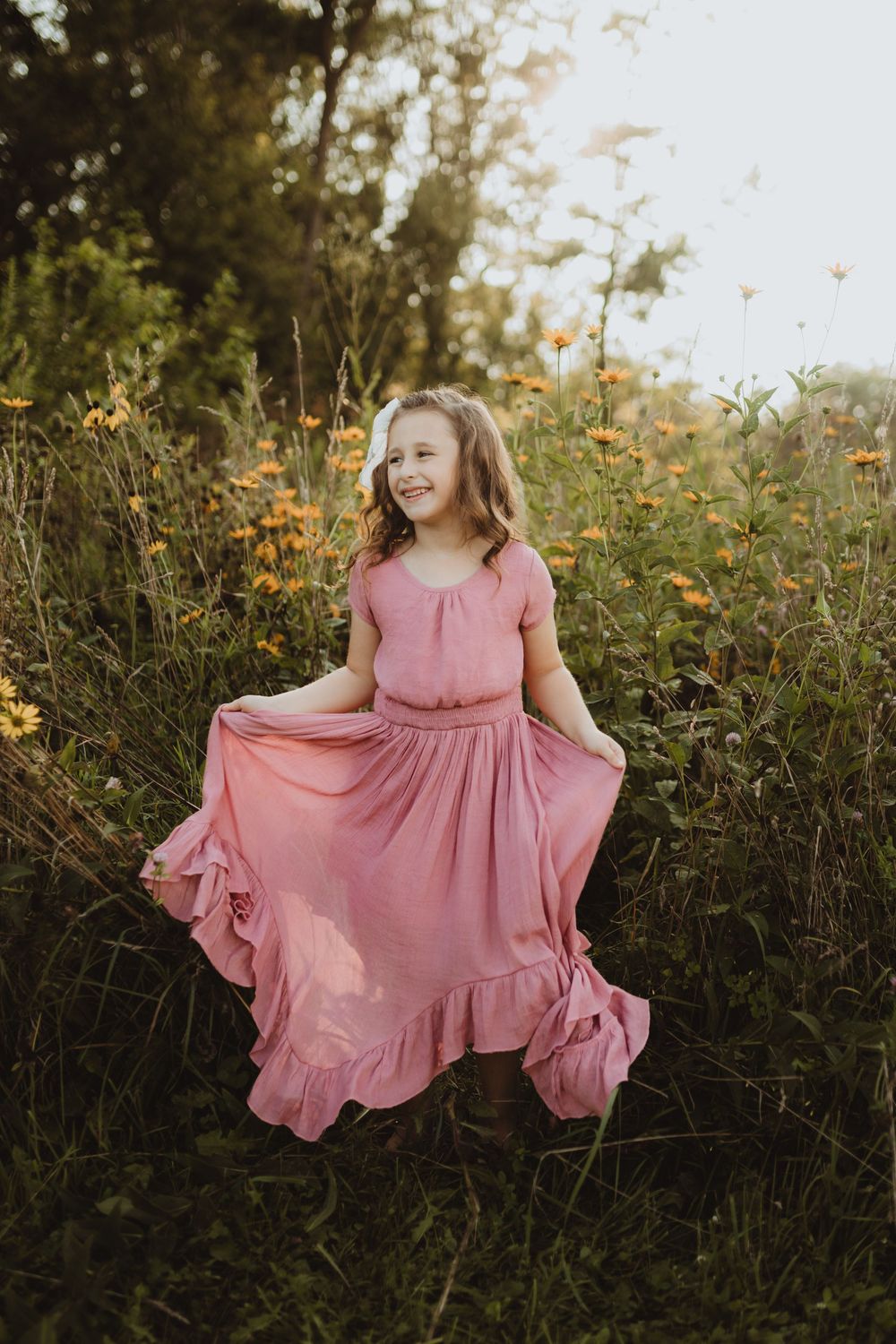 A young girl in a flowing pink ruffled dress twirls among wildflowers in a sunlit garden.