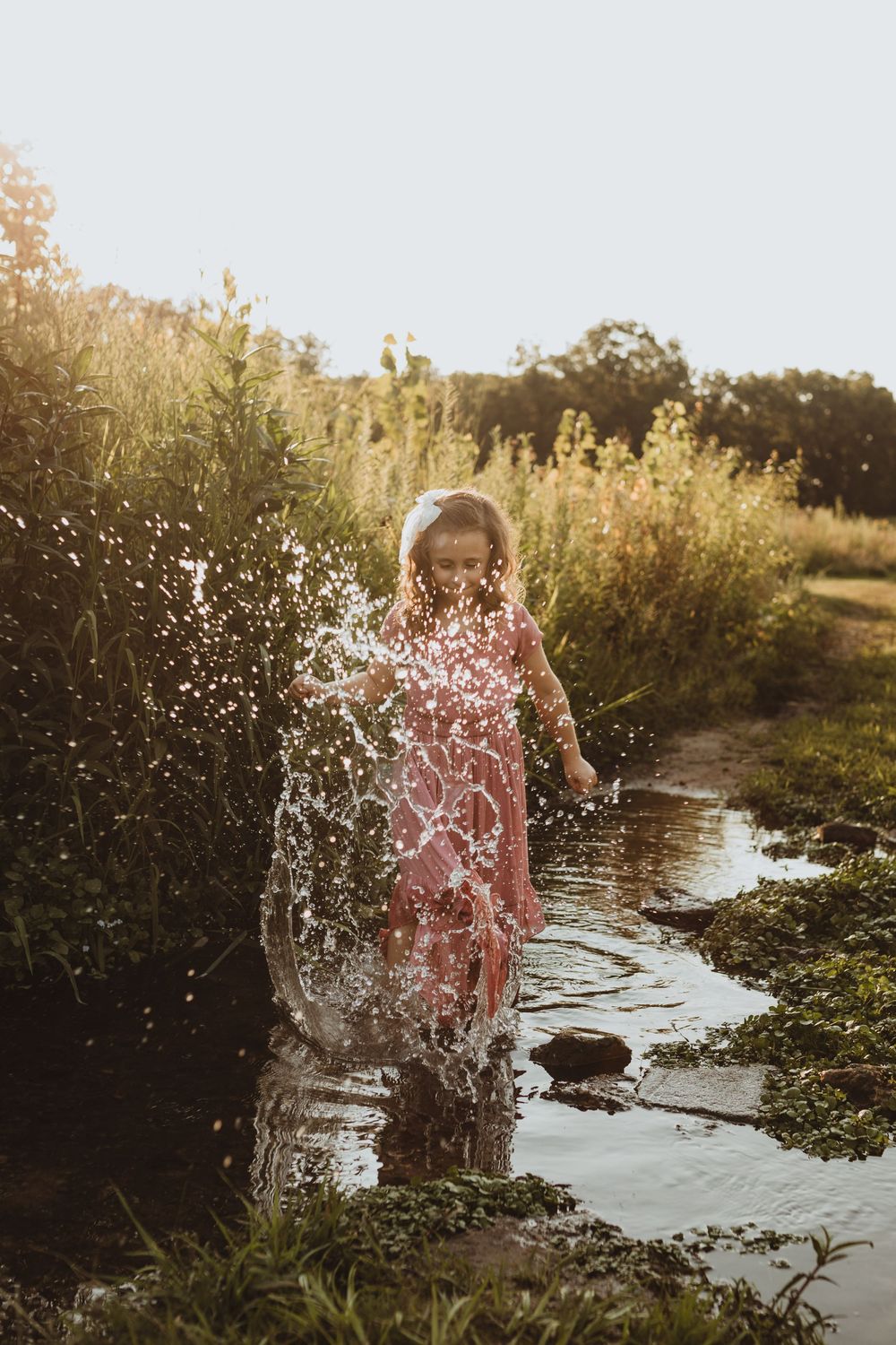 Young girl in pink dress splashing in a puddle outdoors, bathed in golden sunset light.