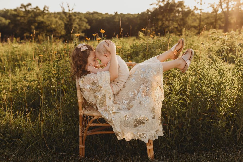 A mother sits on a wooden chair in a sunlit meadow, lovingly embracing her young daughter in a floral lace dress.