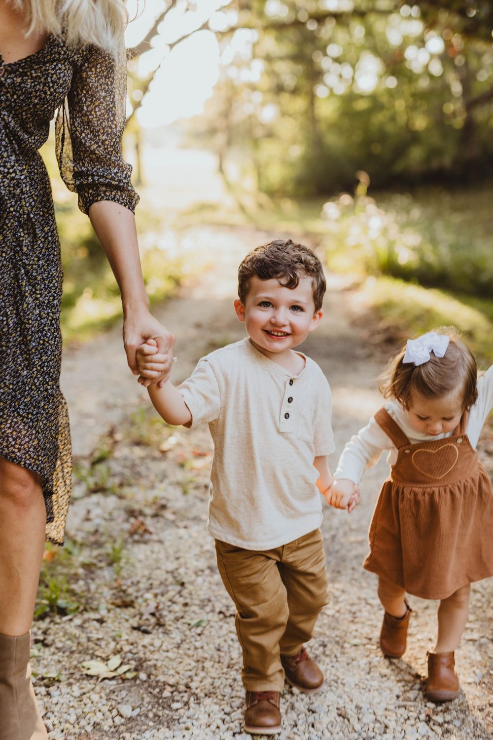 Two young children walk hand-in-hand with a woman on a sunlit path, smiling joyfully during an outdoor family photo session.