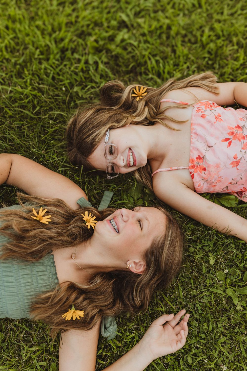 Two smiling girls lying on green grass with yellow flowers in their hair, wearing summer dresses.
