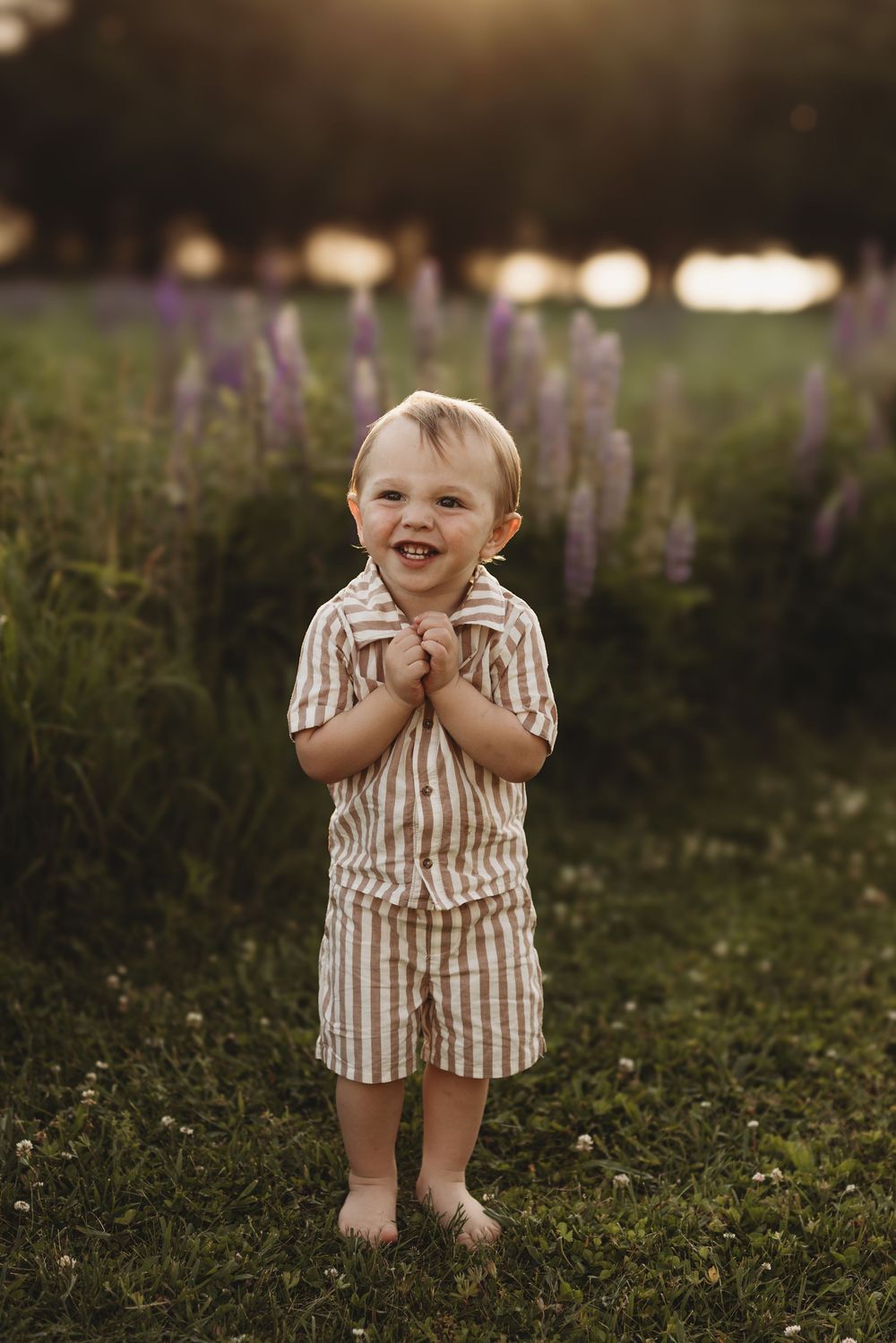 Smiling toddler boy in striped outfit standing barefoot in a field of purple lupine flowers at golden hour.