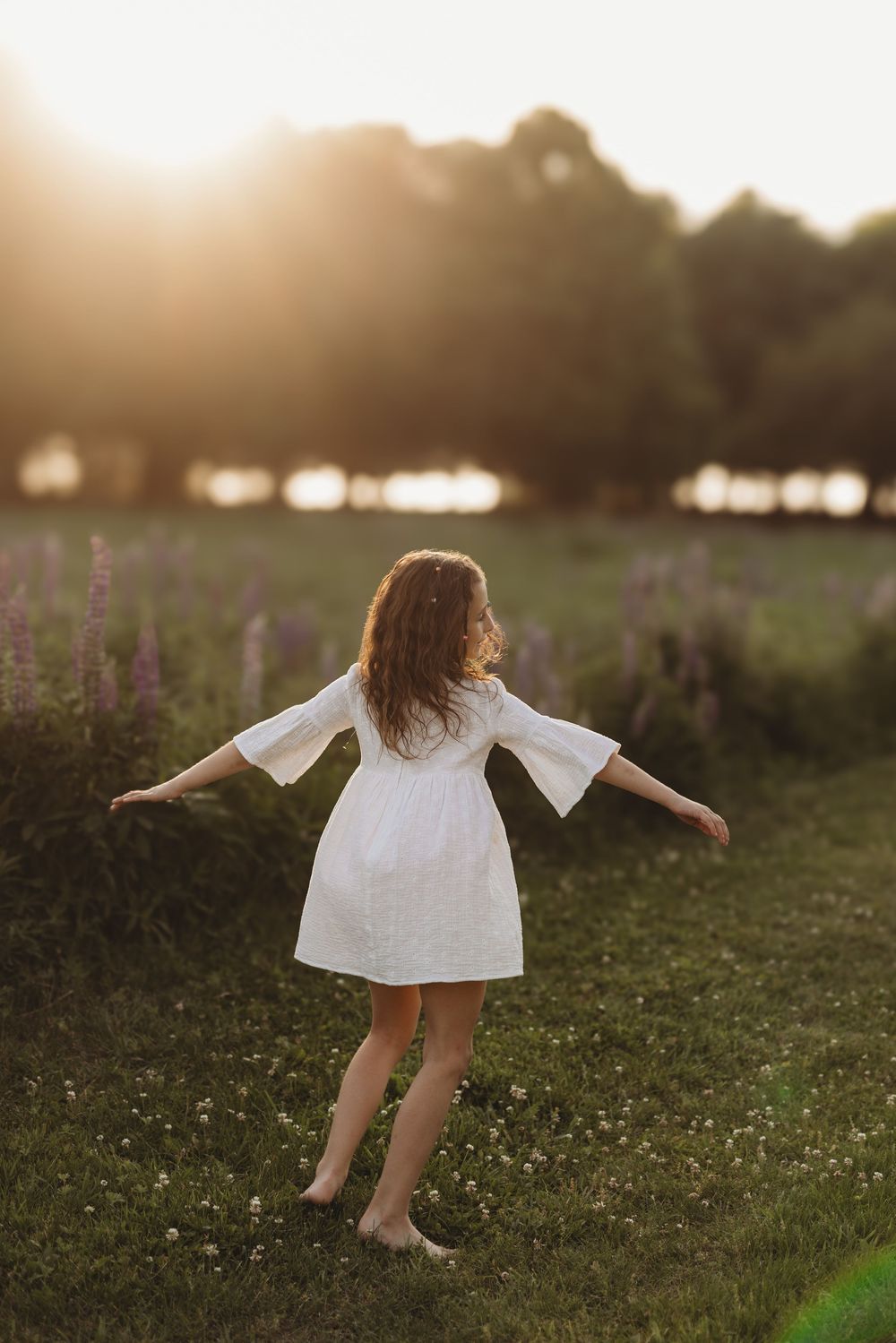 Girl in white dress twirling barefoot in a sunlit meadow with purple wildflowers at golden hour.