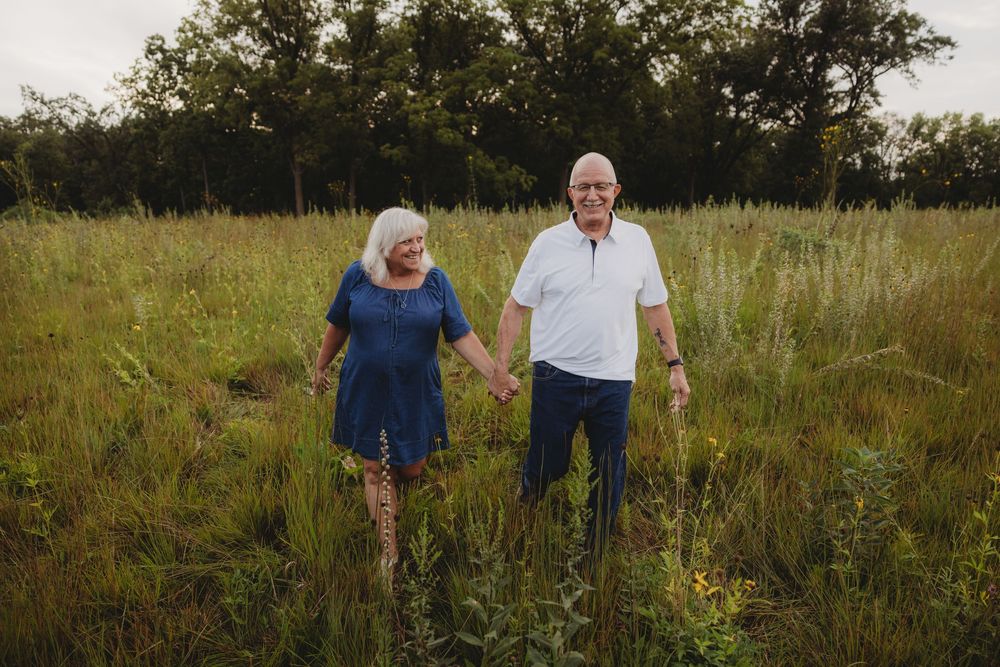 Elderly couple holding hands and walking through a lush green meadow, wearing casual blue and white outfits.