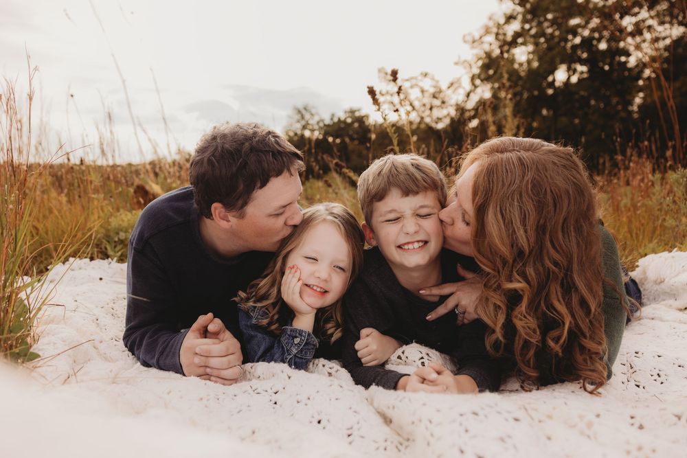 Family of four lying on white blanket outdoors, parents kissing cheeks of two laughing children in golden field.
