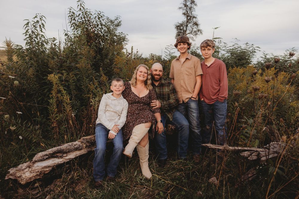 Family of five posing outdoors on a log surrounded by lush greenery in a warm autumn nature setting.