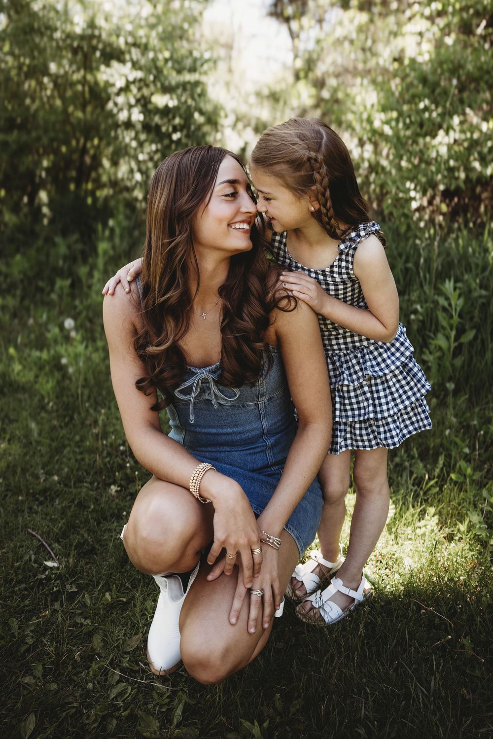 A mother crouches in a green park, smiling at her young daughter in a plaid dress, sharing a tender moment outdoors.