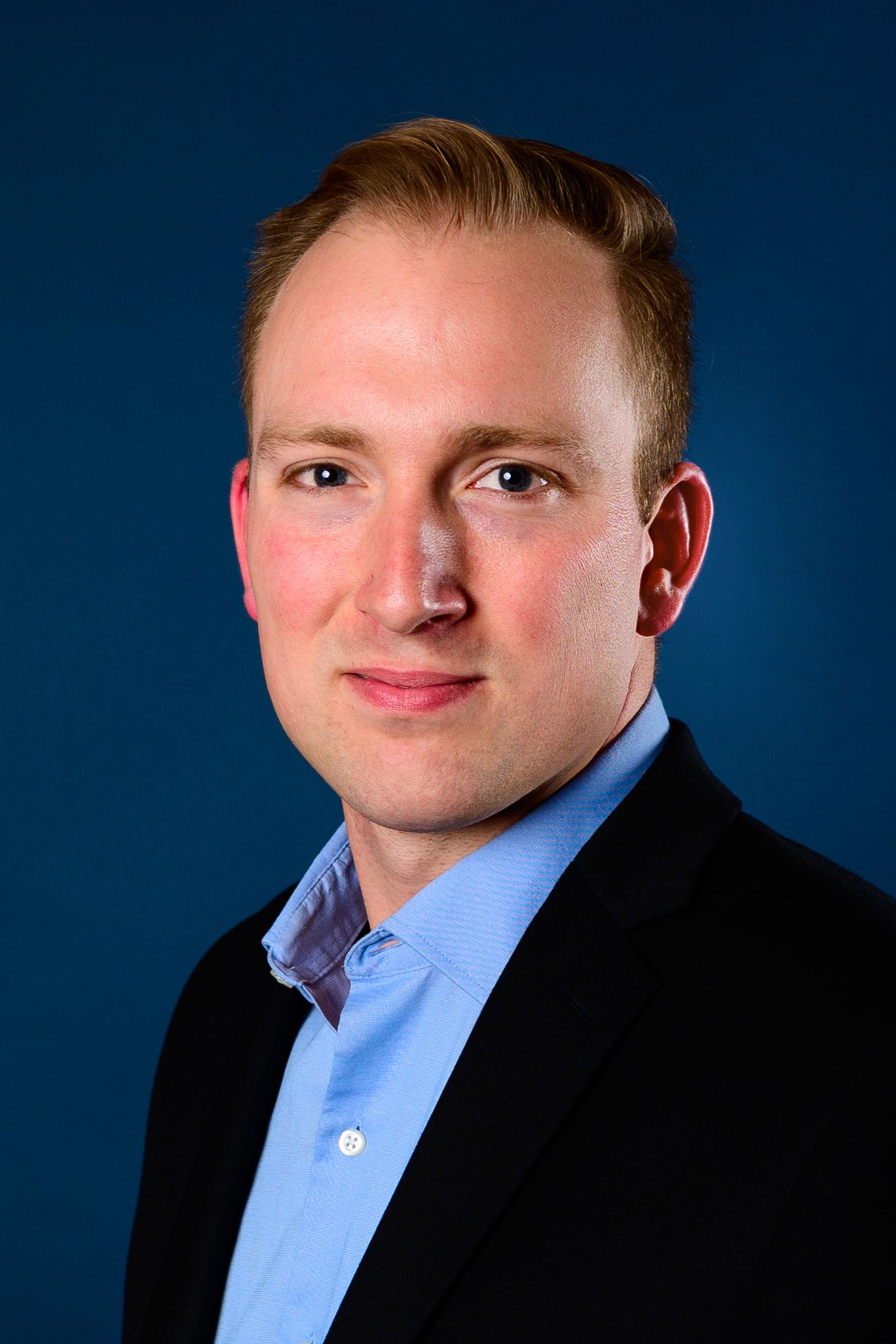 Professional headshot of a young man with red hair wearing a black blazer and light blue shirt against a dark blue background.