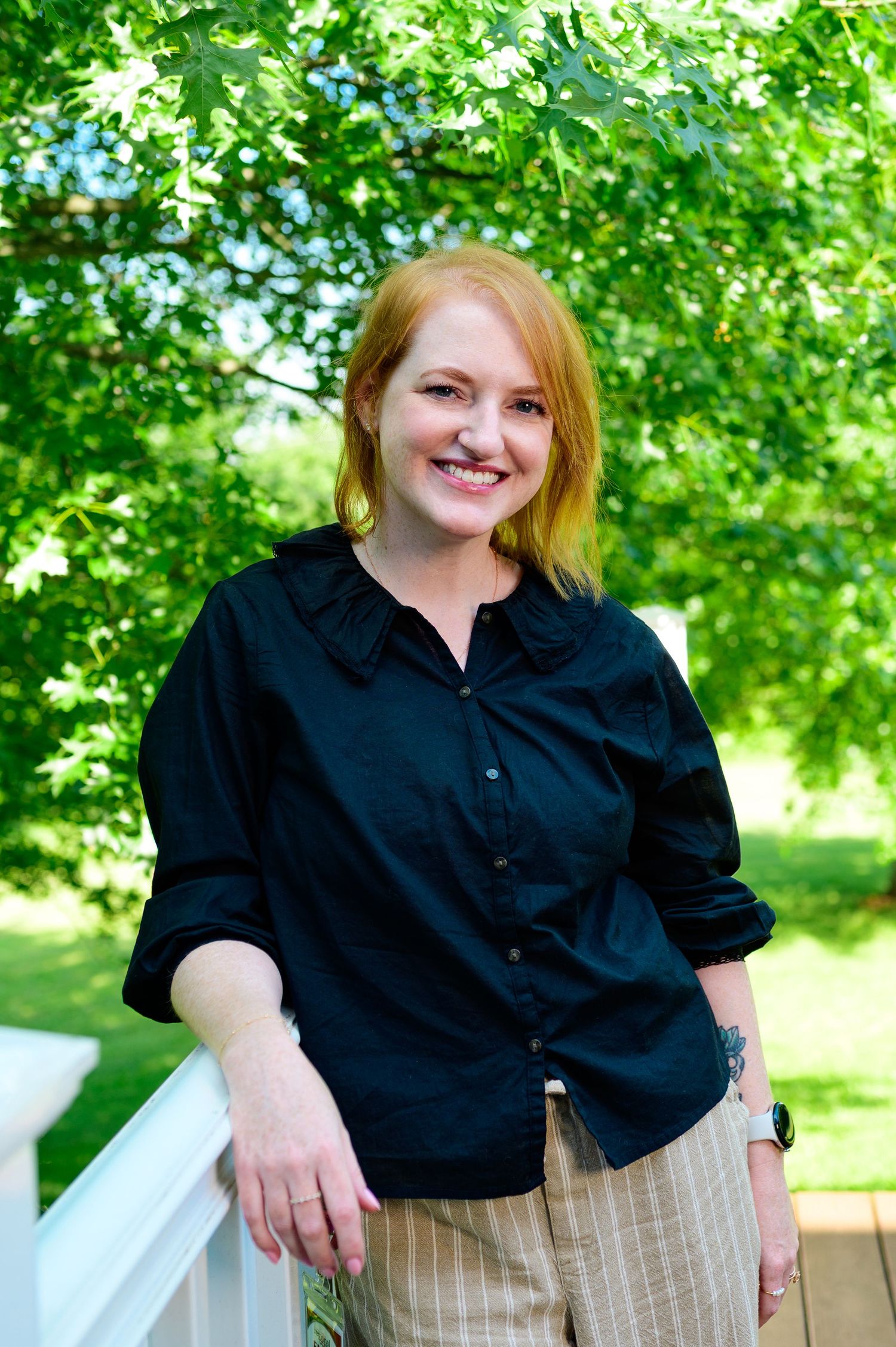 Smiling redhead woman in black blouse and plaid pants standing on white porch surrounded by lush green trees.