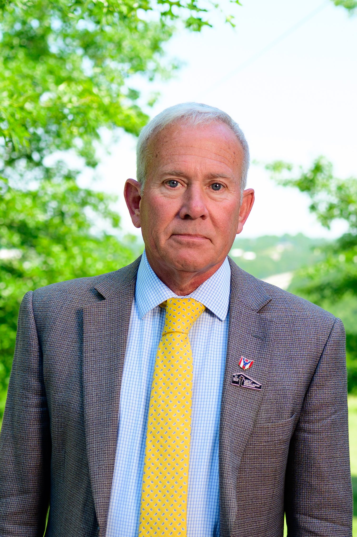 Professional man in gray suit and yellow tie poses outdoors with green trees in background.