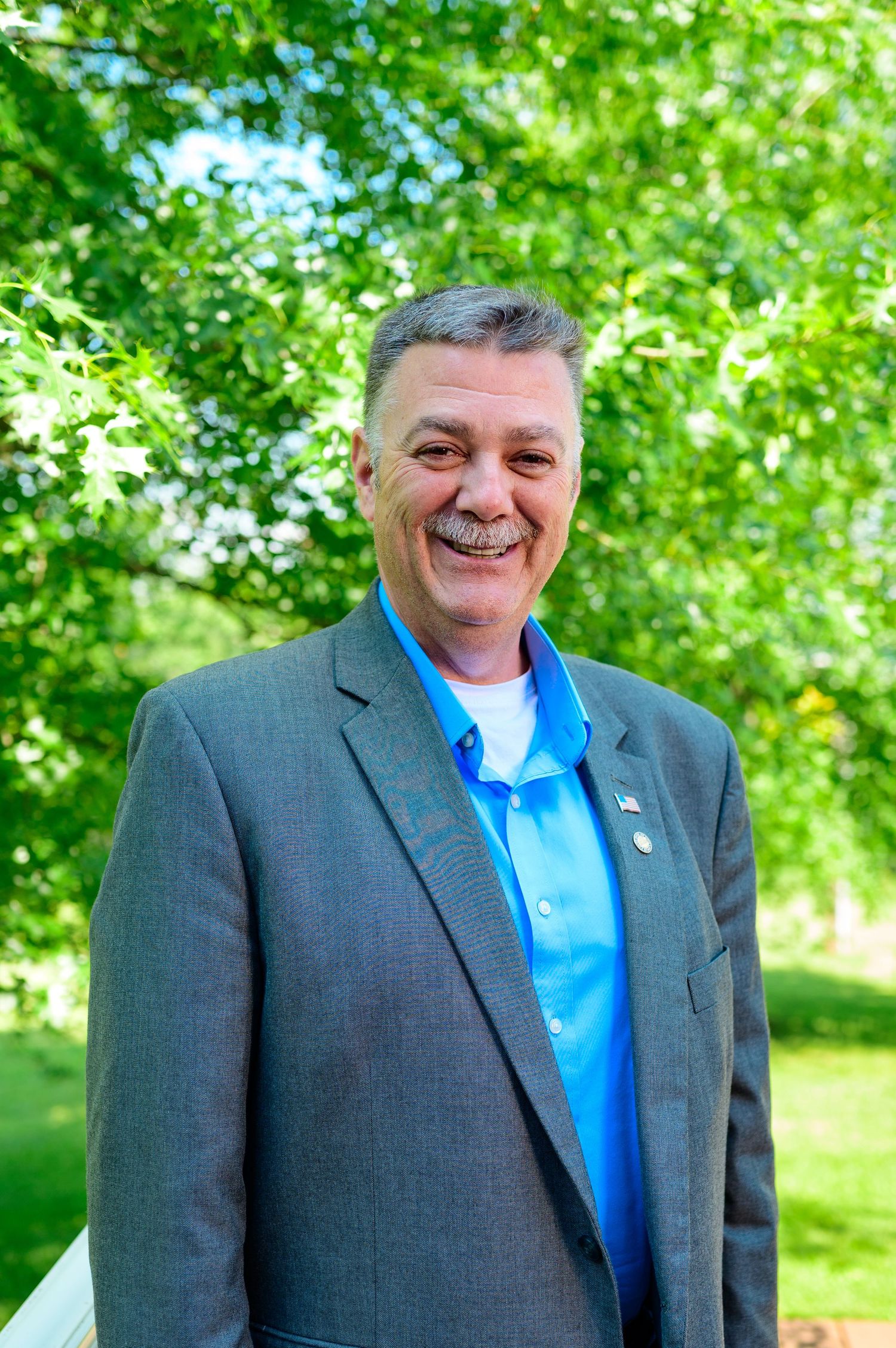 Smiling middle-aged man in gray blazer and blue polo shirt outdoors with green trees in background.