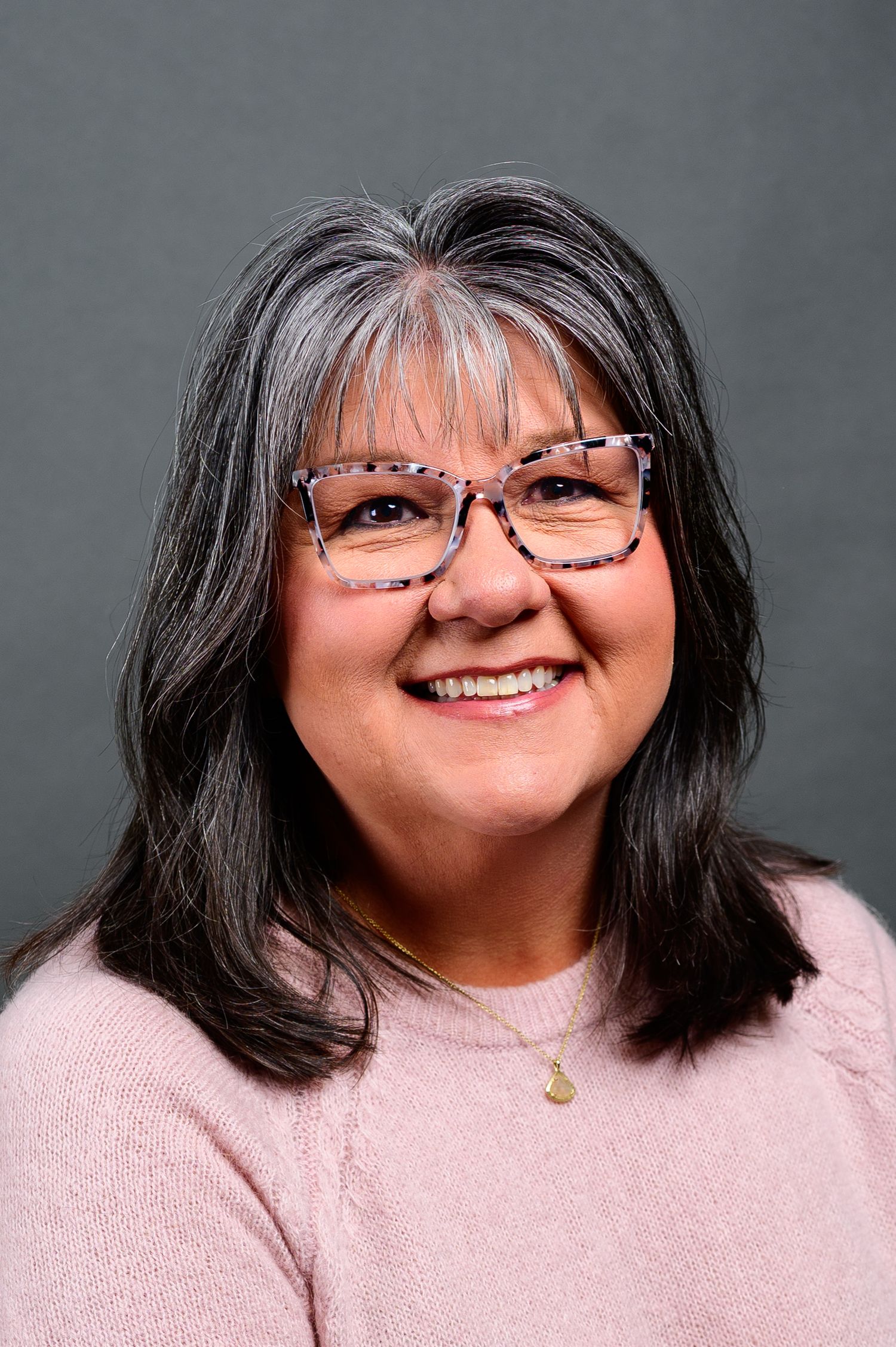 Smiling middle-aged woman with gray-streaked hair, pink glasses, and a light pink sweater poses for a professional headshot.