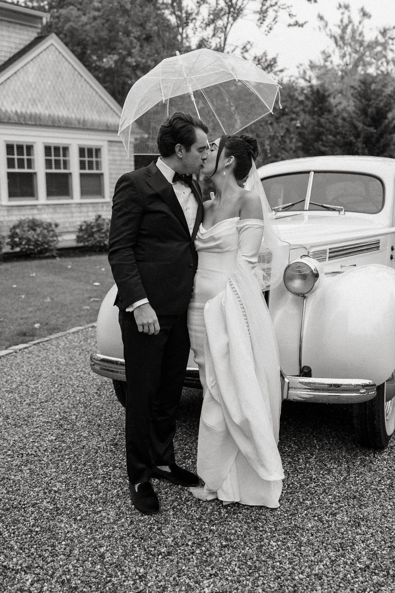 Bride and groom kissing under a clear umbrella beside a classic white vintage car in the rain. Cape Cod, Ma.  Miranda Photo & Co