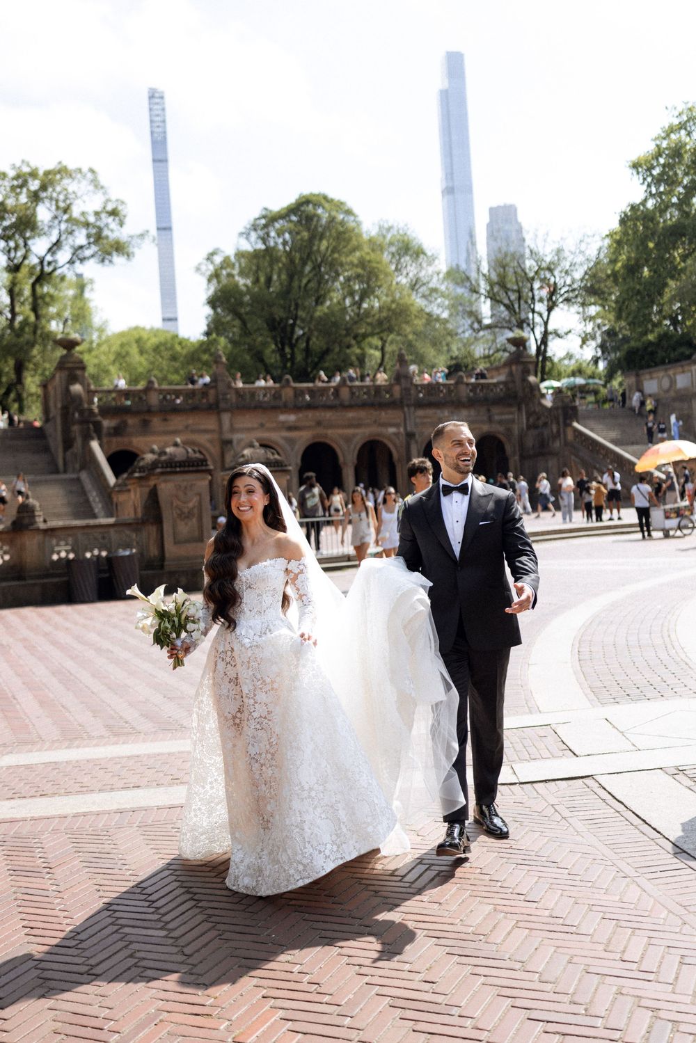 A couple in wedding attire walks along a brick paved path during a sunny outdoor photoshoot.