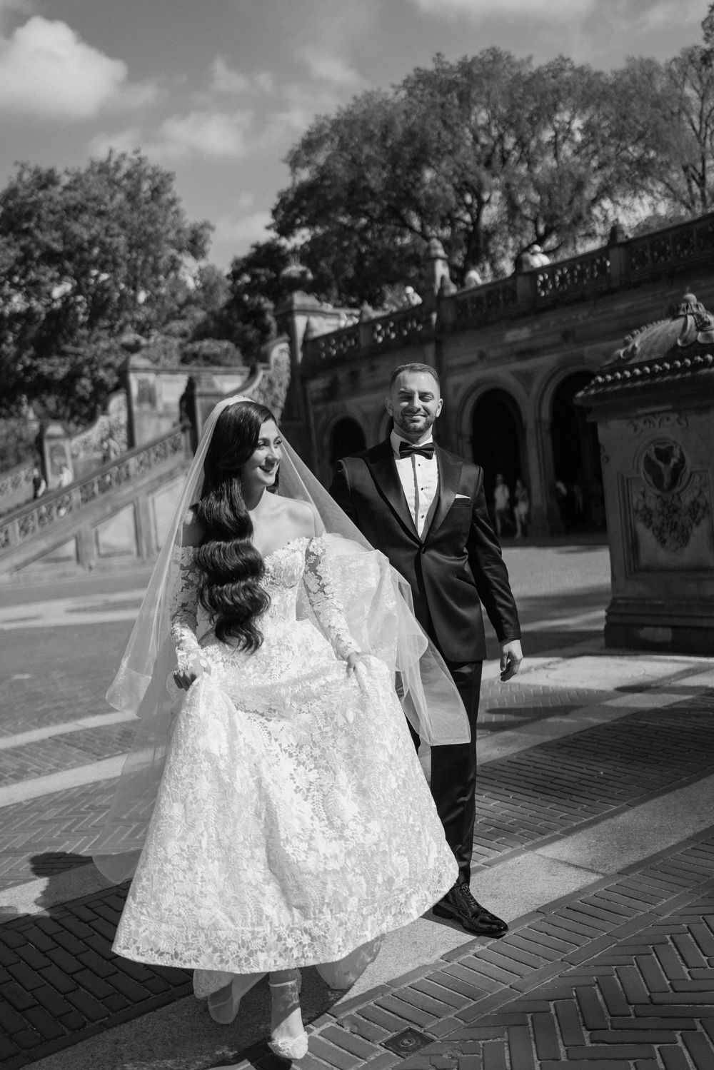 A couple in formal attire walks along a brick pathway during a black and white photoshoot.