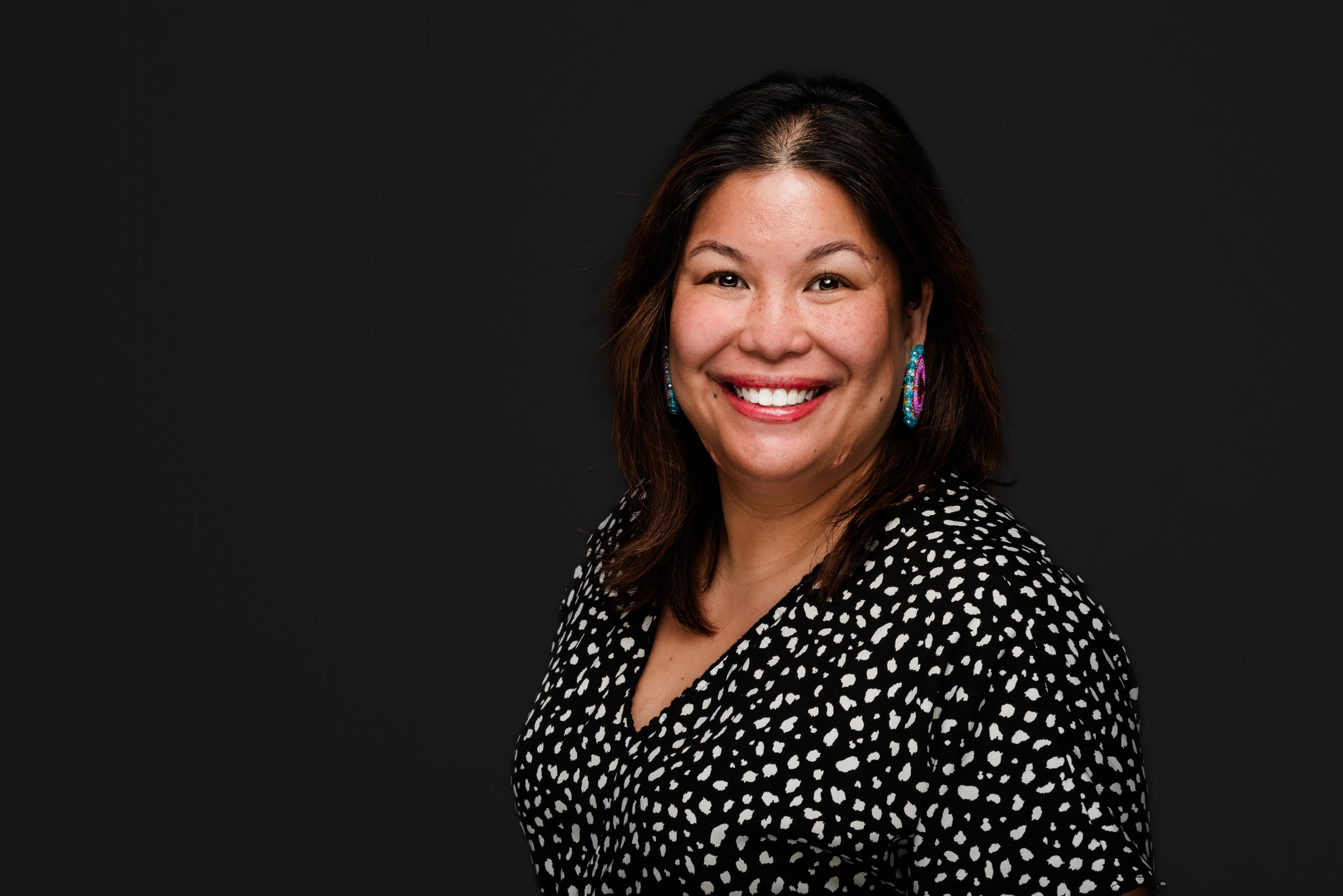 Professional headshot of someone wearing a black and white polka dot top against a dark background.