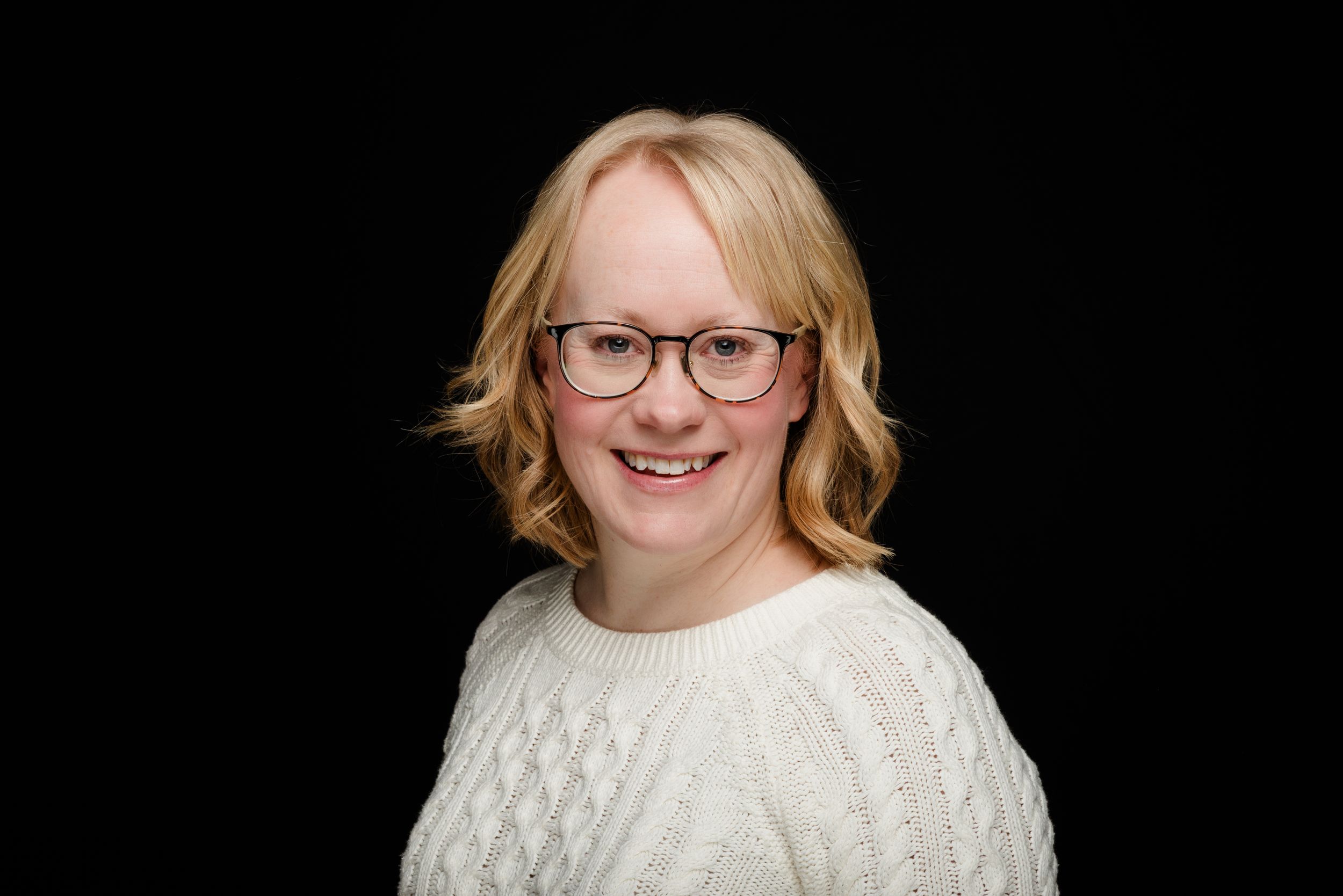 Professional headshot of a person in glasses and white cable knit sweater against a dark background.