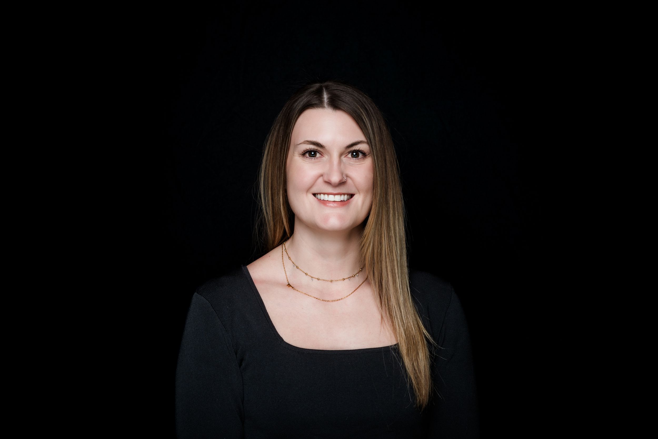 Professional portrait of a person with long brown hair wearing a black top and necklace against a dark background.