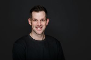 A person wearing a black sweater smiles warmly against a dark background in a professional portrait setting.