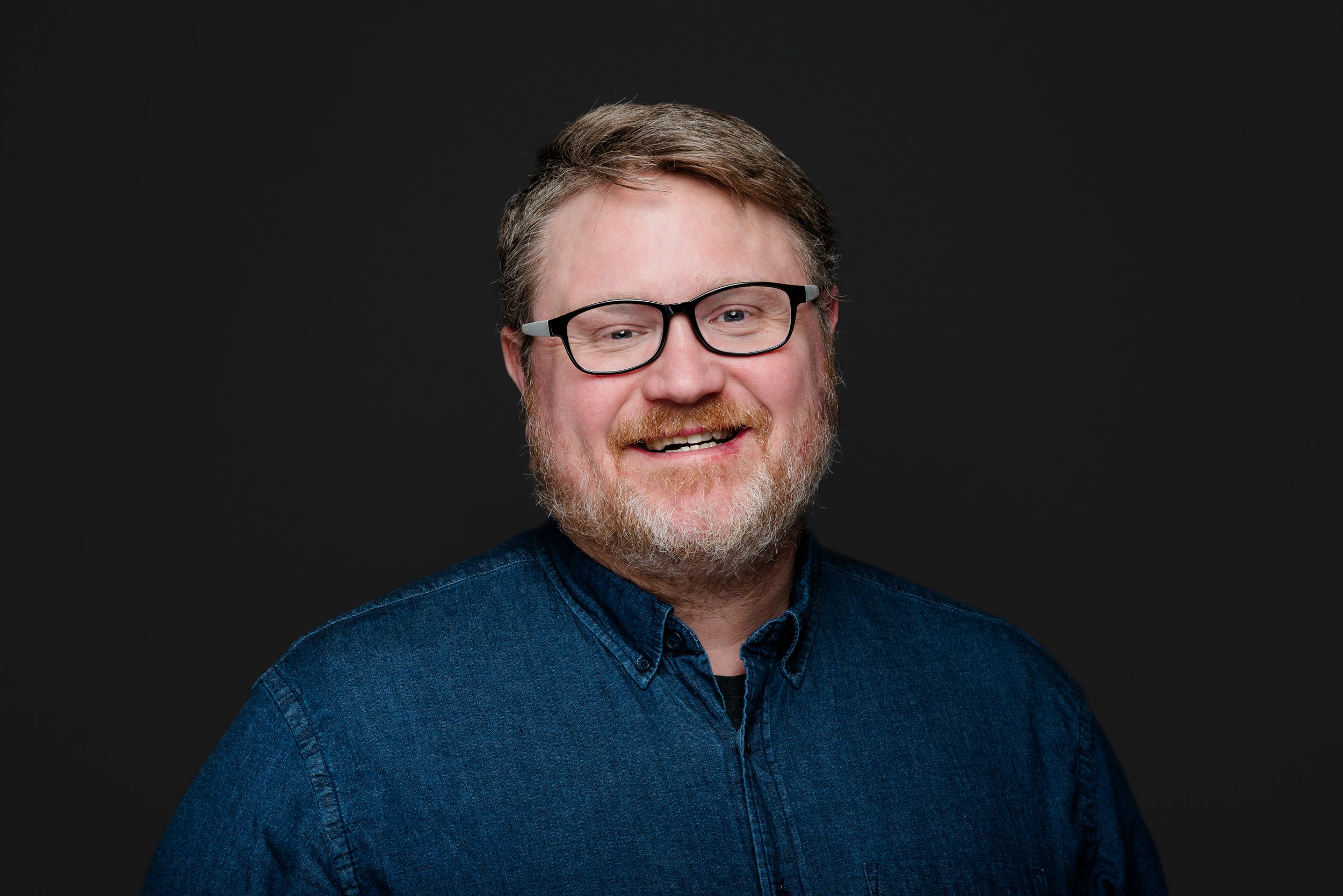 Professional headshot of person wearing glasses and blue button-down shirt against dark background.