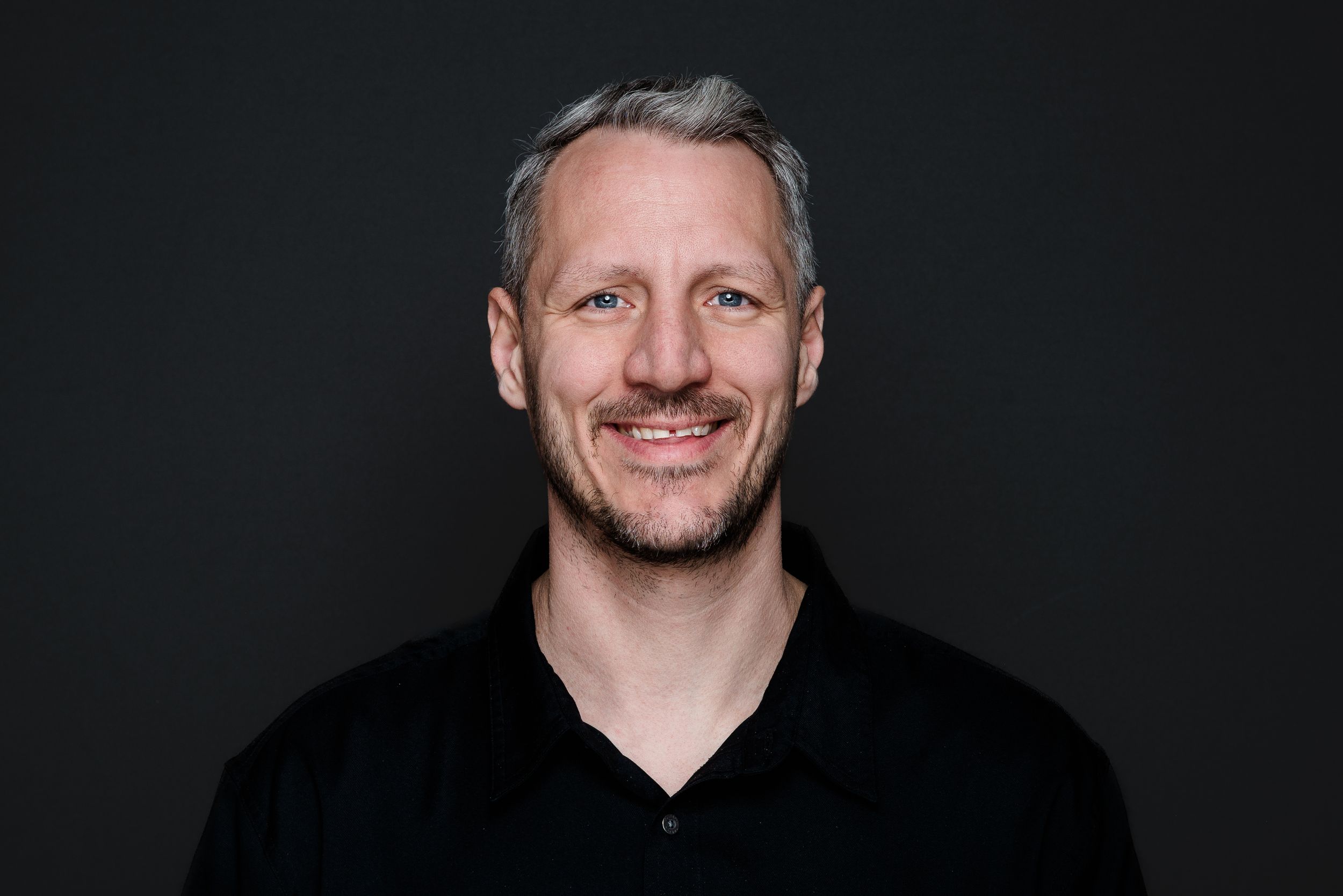 A smiling person wearing a black v-neck shirt poses against a dark background in a headshot portrait.