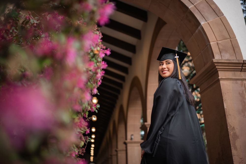 Dallas senior graduation portrait of smiling Latina graduate in black cap and gown under stone archway with pink flowers, Class of 2026