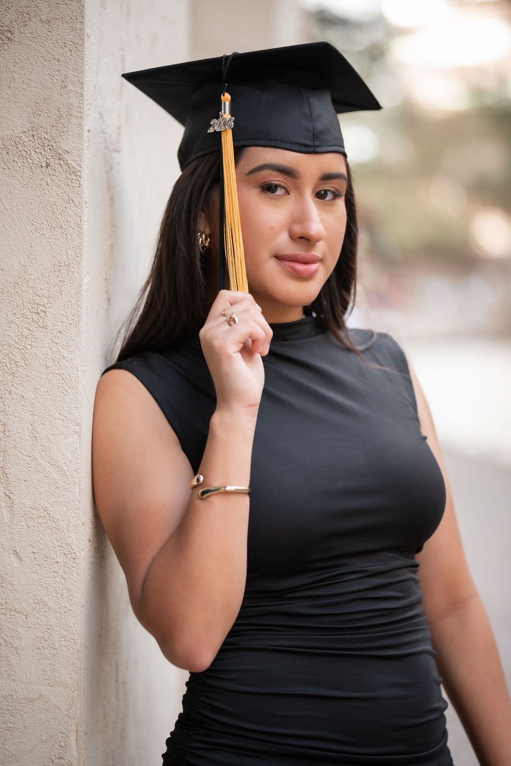 Young woman in black dress wearing a graduation cap with gold tassel, posing outdoors against a wall.