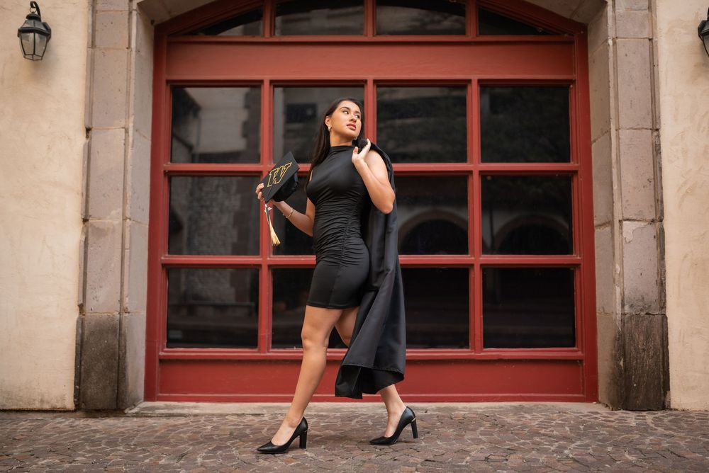 Dallas senior graduation photo of stylish graduate in black dress and heels holding cap and gown, posing in front of red window, Class of 20