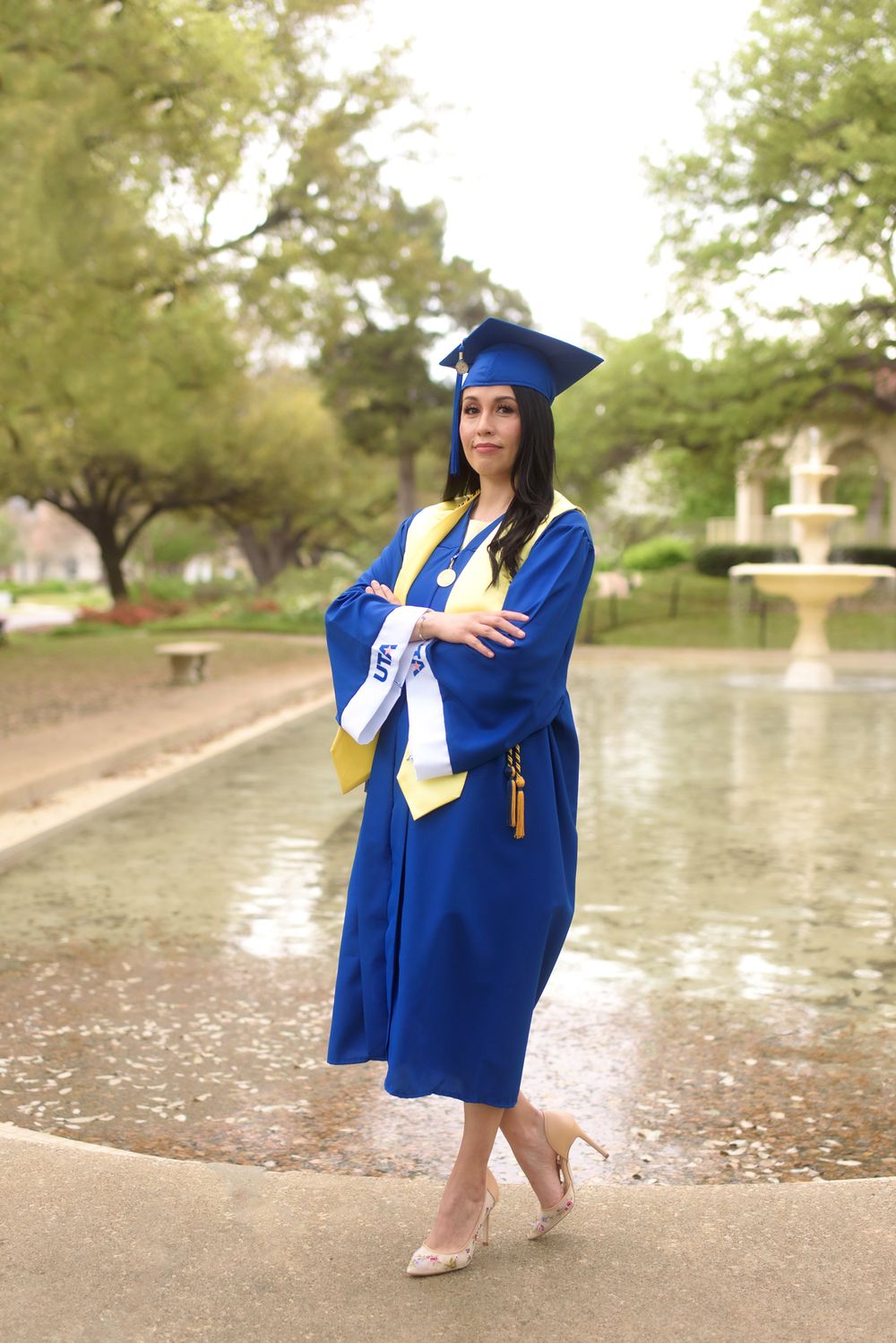 University of Texas Arlington Nursing Graduate in Cap and Gown posing in front of fountain.