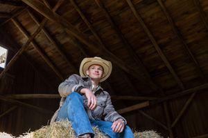 A farmer in a straw hat gathers hay in a rustic wooden barn loft on a sunny afternoon.