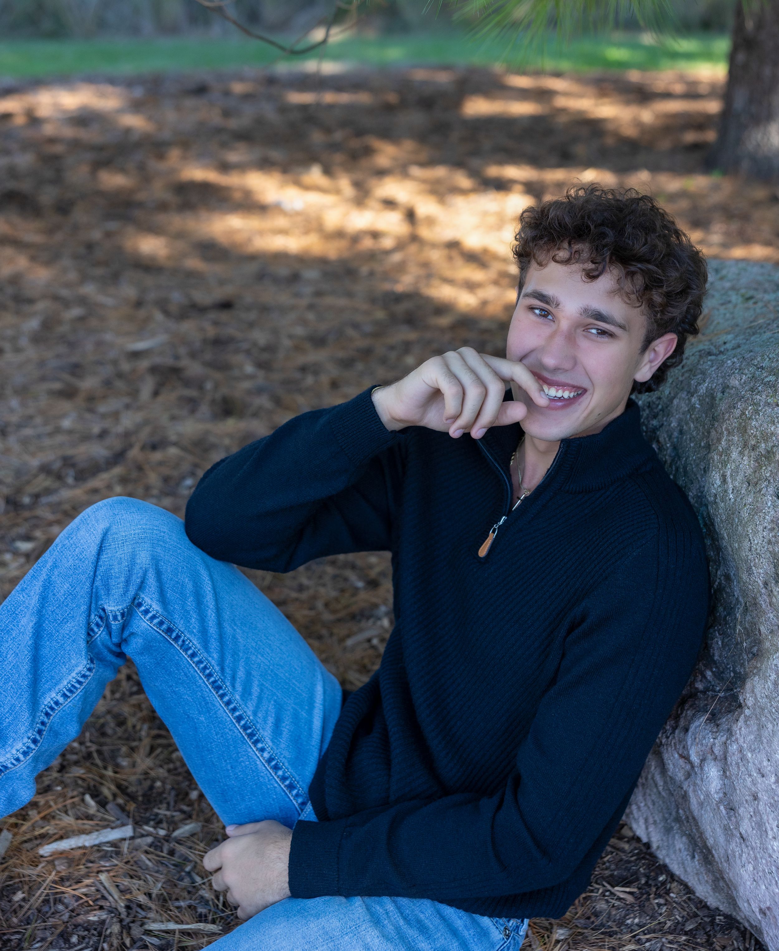 A person in a black sweater and jeans relaxes against a tree trunk in dappled sunlight on a forest floor.