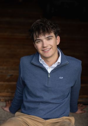 Portrait of someone wearing a blue quarter-zip sweater and white collared shirt against a dark wooden background.