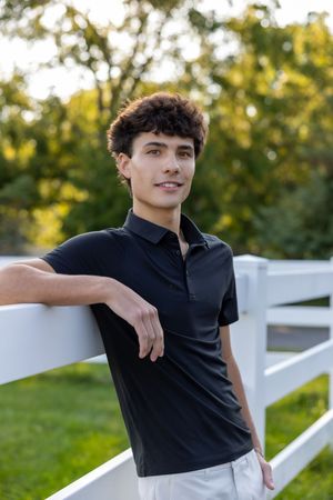Person in black polo shirt poses casually against white fence in outdoor setting with green foliage background.