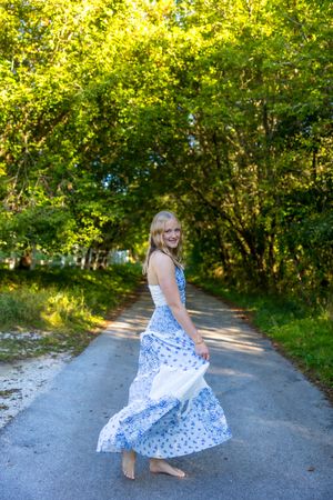 A person in a blue and white polka dot dress twirls playfully on a tree-lined path on a sunny summer day.