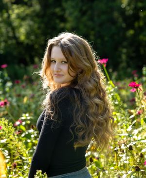 A person with long wavy hair poses in a garden with vibrant red flowers and lush greenery in the background.