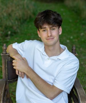Young person in a white polo shirt poses outdoors on a wooden chair with ornate details against a green background.