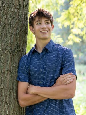 Young person in a blue polka dot shirt poses casually against a tree trunk with natural sunlit foliage in background.