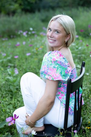 Someone wearing a floral print top and white pants sits on a black chair in a garden with pink wildflowers.