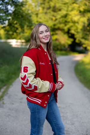 A varsity letterman jacket in red and cream colors styled with blue jeans on a gravel path with autumn trees in background.