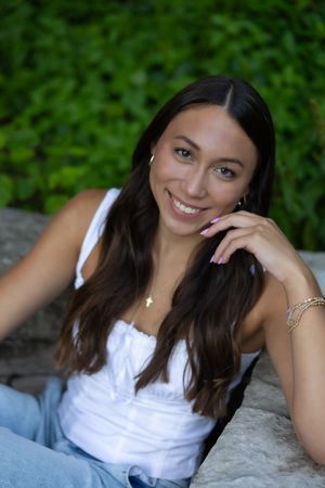 A smiling person in a white tank top and jeans sits on rocks with greenery in the background.