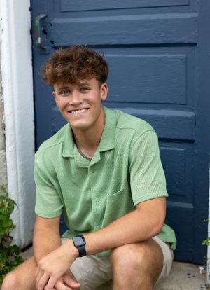 Person in green polo shirt sitting outdoors against blue door smiling at camera.