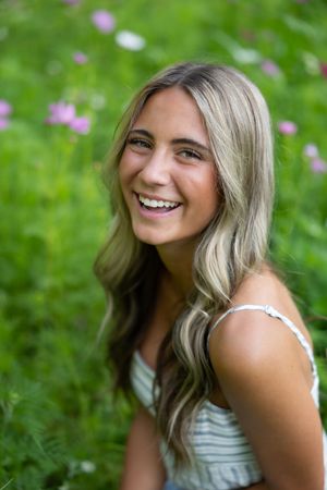 A person with blonde wavy hair poses outdoors in a white top against a green background with purple flowers.