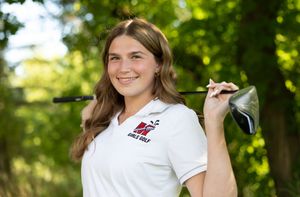 High school golfer in white team polo holds golf club over shoulder while smiling outdoors in sunny setting.