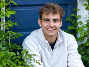 A person in a white sweatshirt smiles while sitting outdoors against a blue wall with green climbing vines.