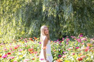 A person in a white dress stands among vibrant wildflowers in a sunny garden setting with willow trees in the background.