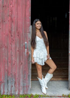 A person in a white top and shorts poses in front of a rustic red barn door in a casual outdoor photoshoot.