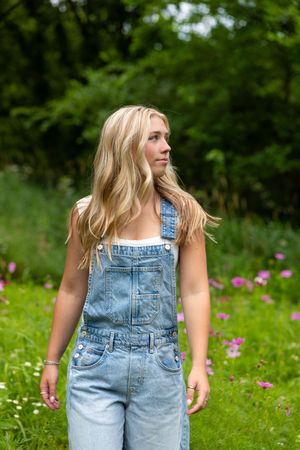 A person wearing denim overalls stands in a green meadow with blooming wildflowers on a sunny summer day.