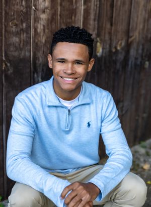 Young person in light blue quarter-zip sweater with small logo sits outdoors against rustic wooden wall background.