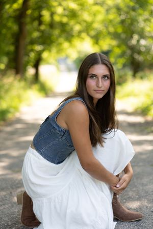 A series of outdoor portrait photos showing someone in a white dress and boots sitting on a natural path in dappled sunlight.