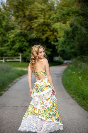 A person in a floral summer dress twirls playfully on a tree-lined gravel path on a sunny day.